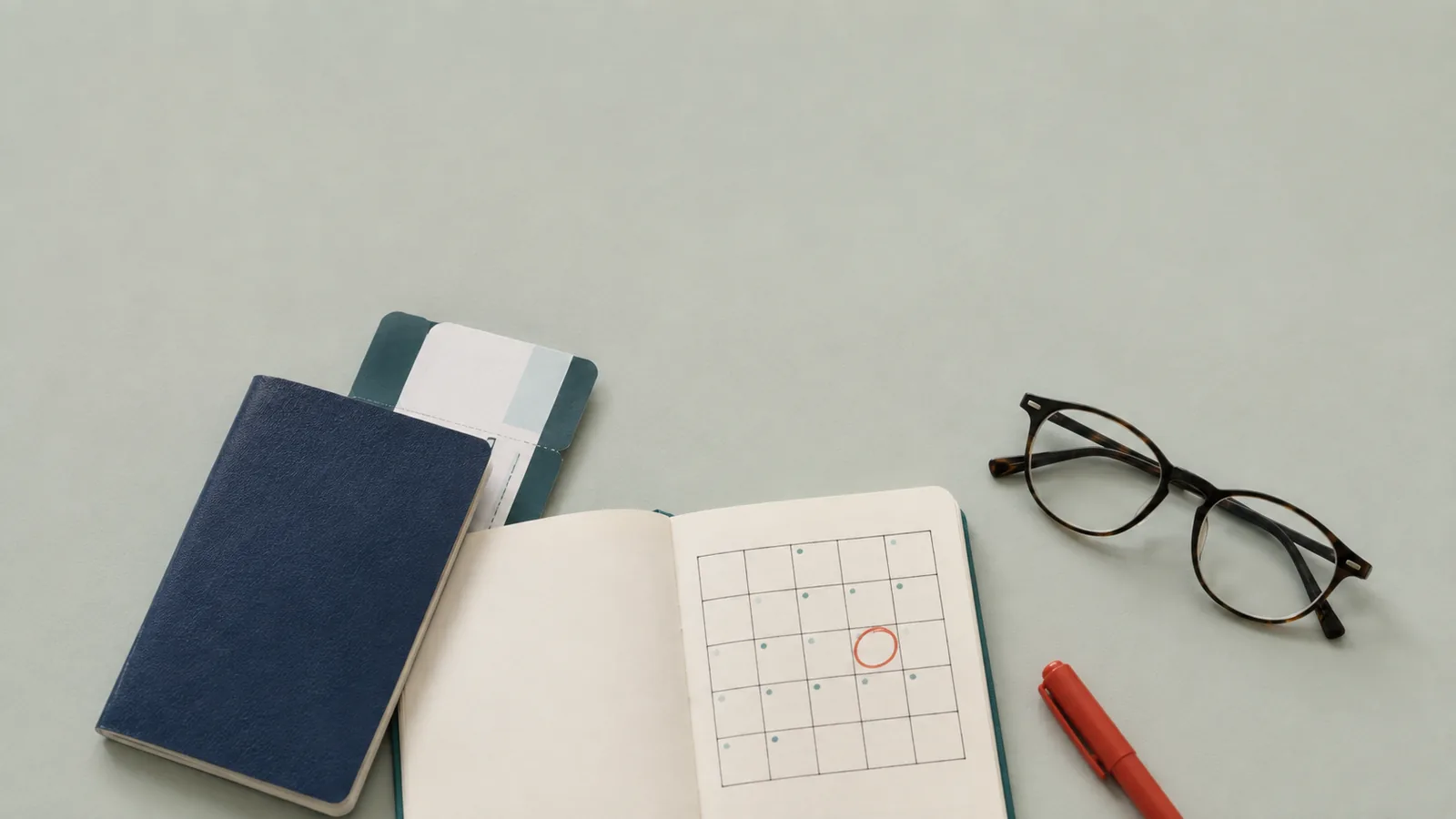 A passport, a boarding pass, an open notebook with a hand-drawn calendar and one date circled in red, glasses and a red pen on a pale surface.